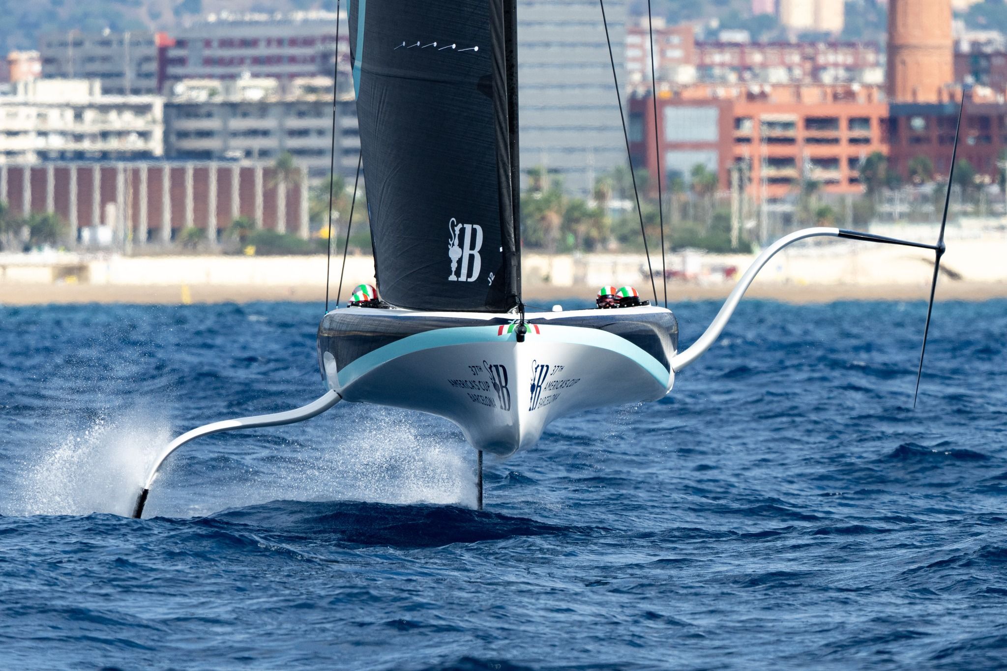 Ein Segelboot mit Tragflächenboot gleitet über das Wasser in der Nähe von Stadtgebäuden im Hintergrund.