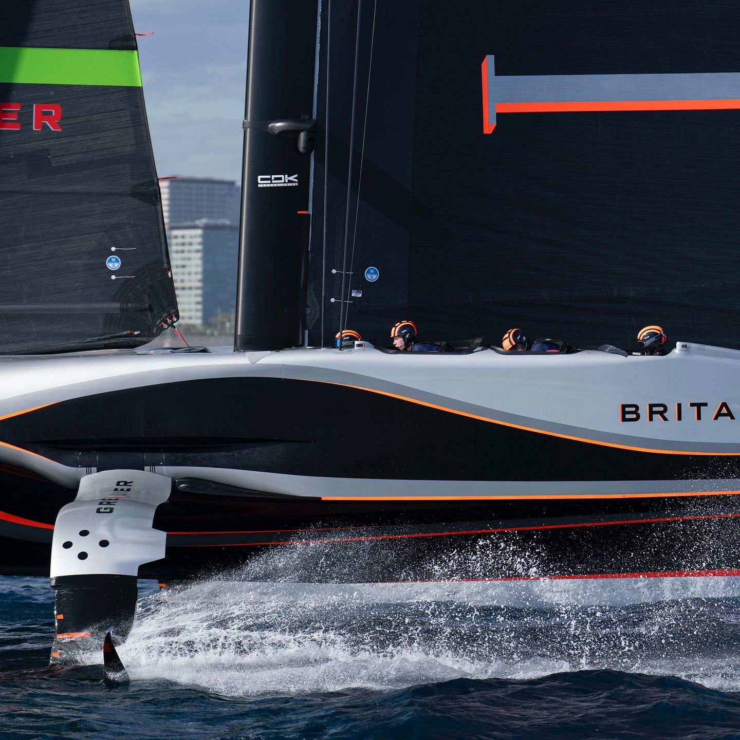 Nahaufnahme des britischen Rennsegelboots, das über dem Wasser gleitet, mit sichtbarer Besatzung und Stadtbild im Hintergrund