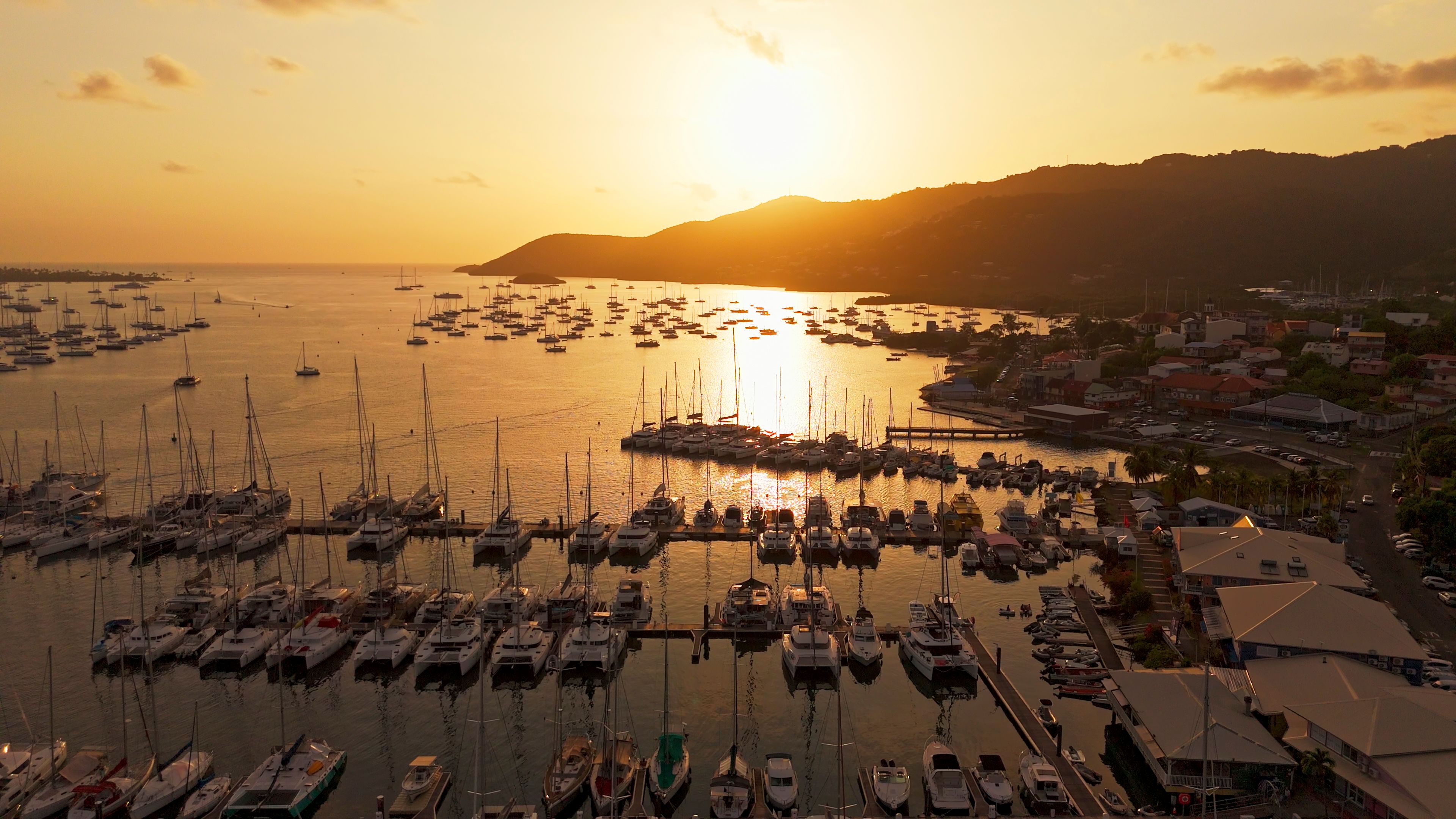 Plachetnice a jachty kotví v klidných vodách martinského přístavu, zatímco slunce zapadá a vrhá na scenérii teplou zlatou záři. V pozadí jsou vidět kopce a pobřežní budovy.