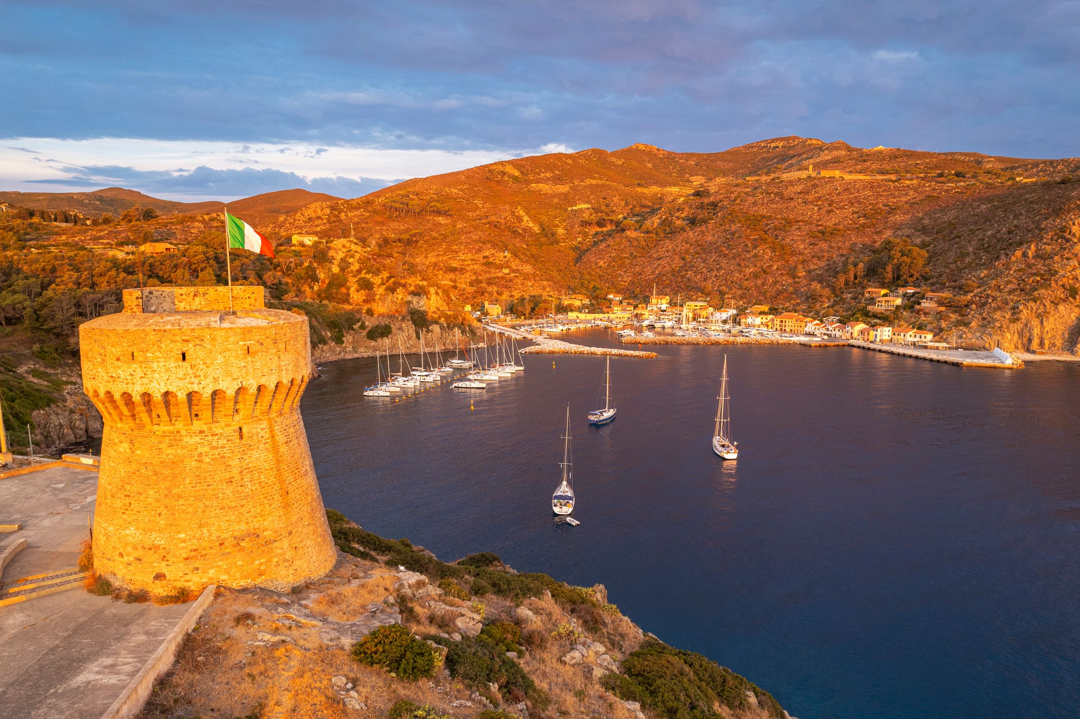 Aerial view at sunrise over the fishing village of Capraia island with the medieval tower in the foreground and the marina in the background, Capraia island, Arcipelago Toscano National Park, Livorno,