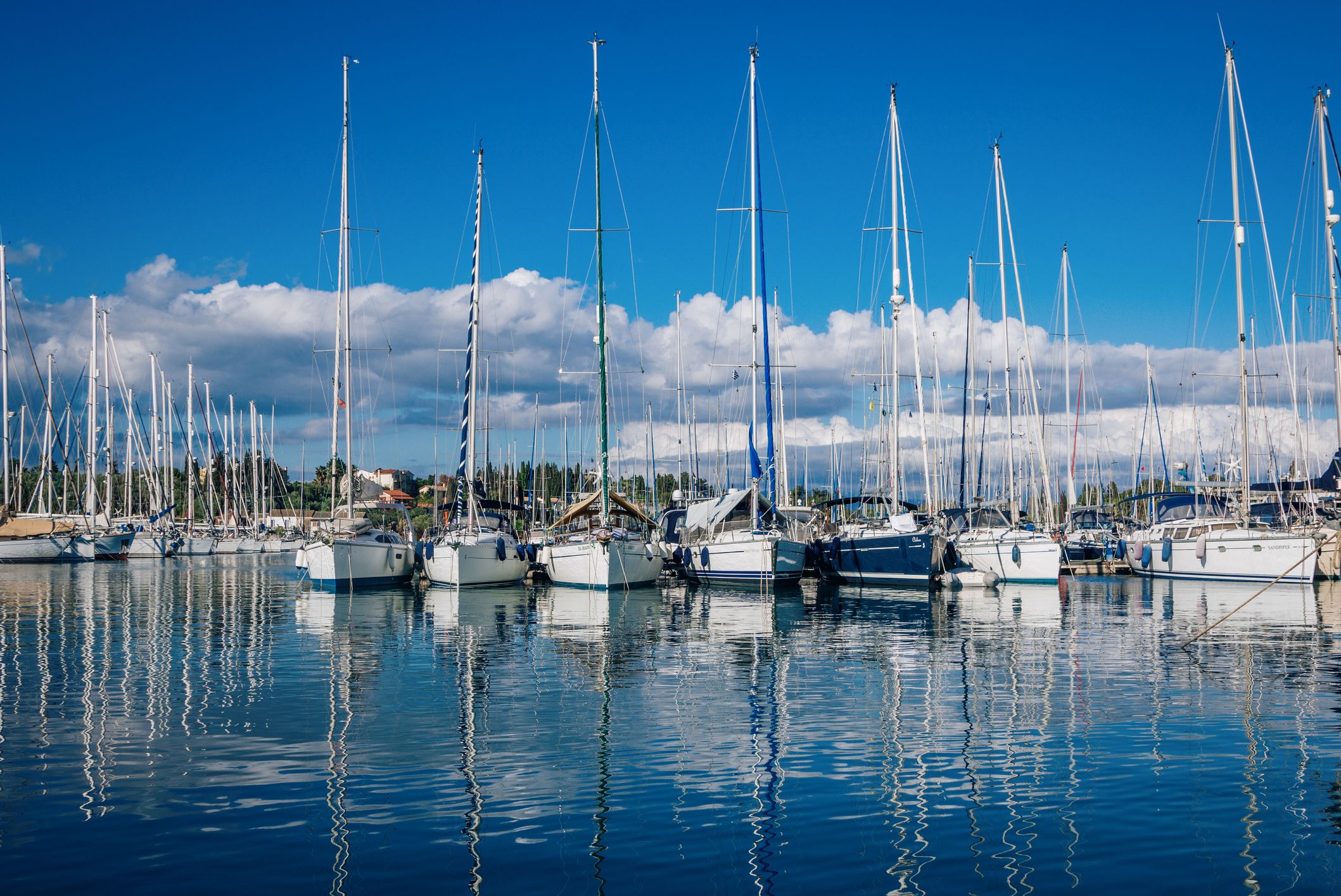 View of beautiful Gouvia port marina - calm sea, white yachts and blue sky with white clouds, reflecting on water surface, and mountains on the horizon.