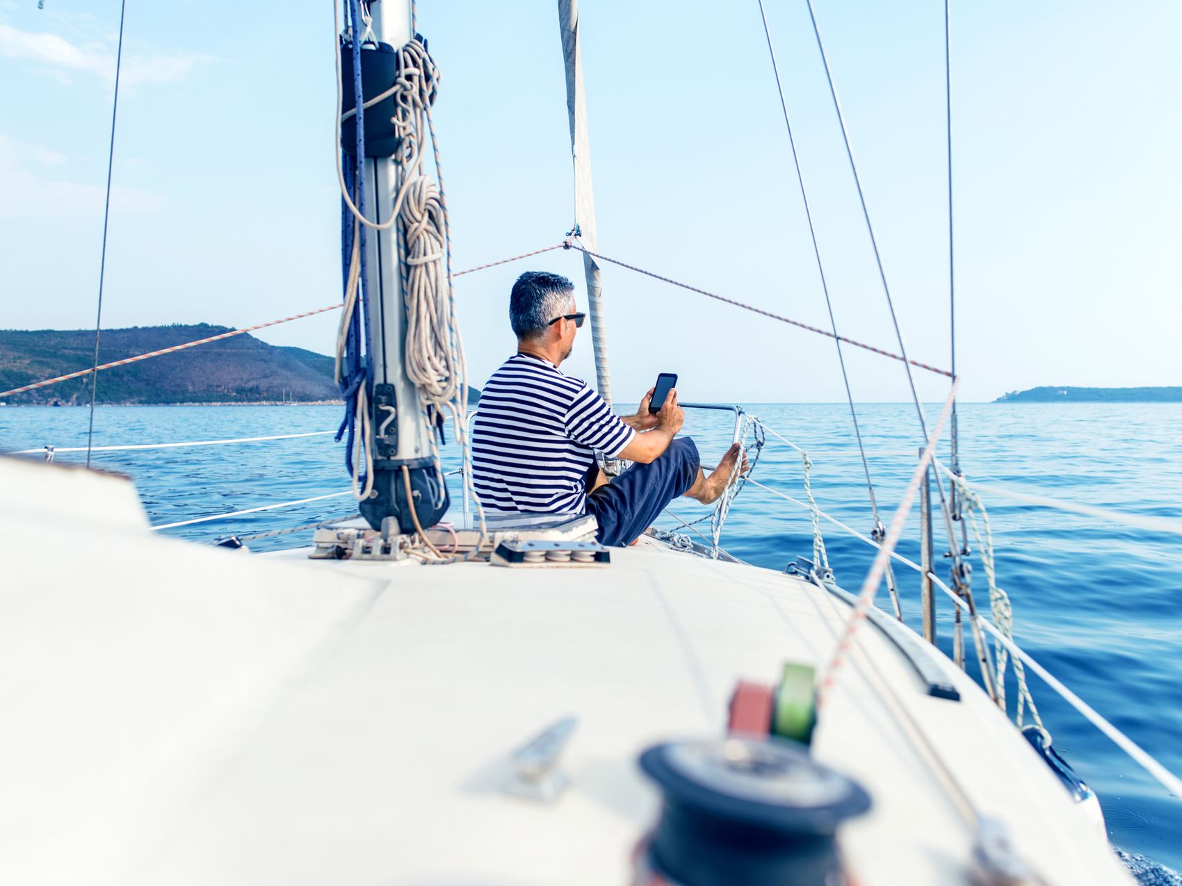 Muž v pruhovaném tričku na jachtě u moře drží telefon